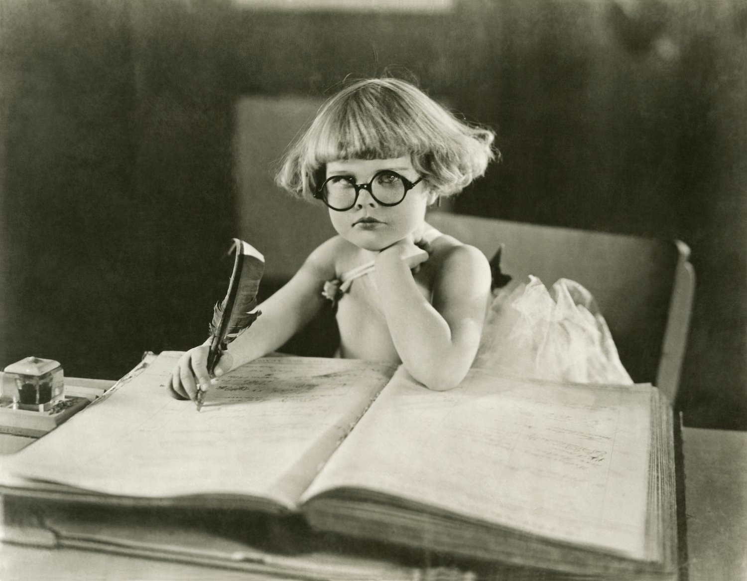 A young child wearing round glasses and writing in a large ledger with a quill pen