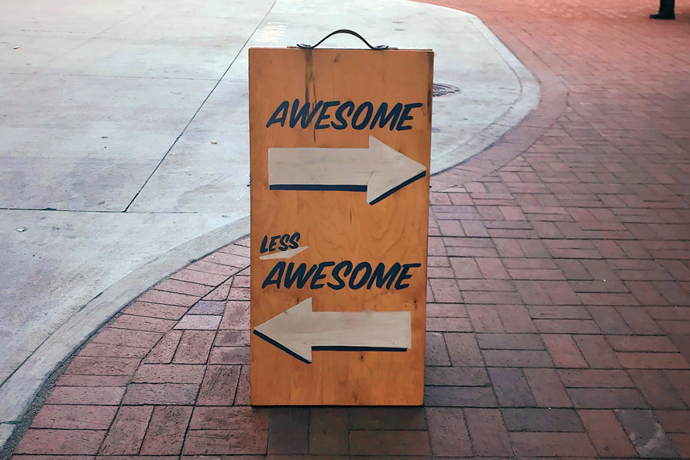 Image: wooden sign on urban sidewalk, painted with two arrows; the arrow pointing right is labeled 'Awesome' and the arrow pointing left is labeled 'Less Awesome'