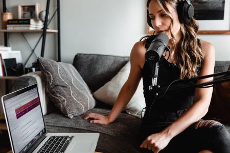 Image: woman on sofa, speaking into microphone while looking at laptop computer.