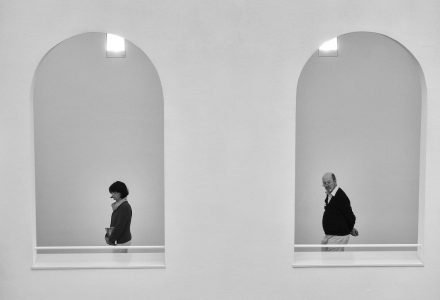 Image: two opens windows in a wall, one frames a woman walking past and the other frames a man walking behind her.
