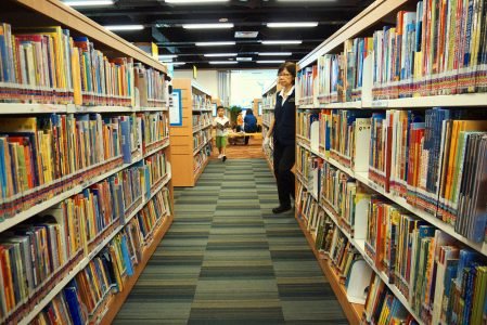 Image: an aisle at a public library, flanked by shelves of books.