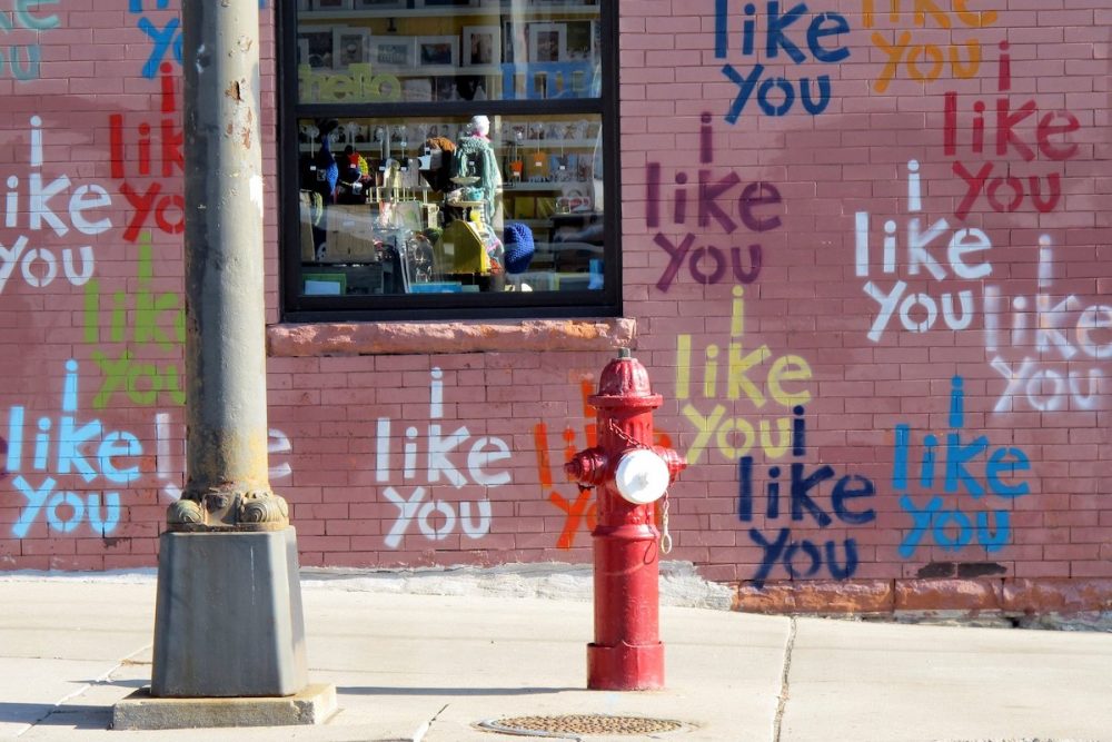 Image: an urban brick wall on which are painted many colorful versions of the phrase "I like you".