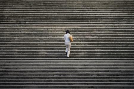 Image: a lone woman walks up an enormous set of stairs which extend off all sides of the frame.