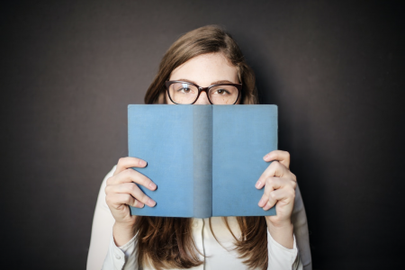 Image: a young woman peers at the viewer over the edge of an open book with no cover design.