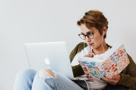 A seated woman wearing earbuds has a pen in one hand and a notebook in the other while she stares intently at an open laptop computer perched on her legs.