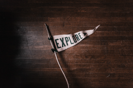 Image: a triangular white flag emblazoned with the word "Explore" is attached to a stick and lying on a dark wooden floor.