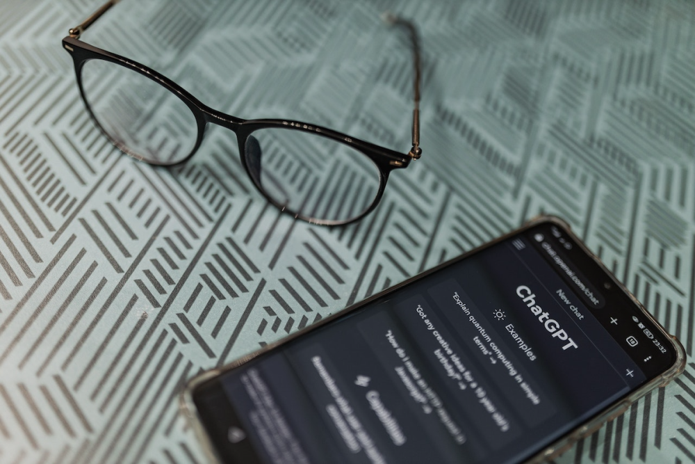 Image: a pair of eyeglasses rests on a table next to a smartphone which is displaying a ChatGPT interface.