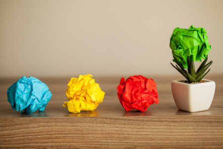Image: three balls of crumpled paper—blue, yellow and red—are in a row at the edge of a desk. Next to them, a fourth crumpled green paper ball rests atop a small potted succulent plant, the effect resembling a tiny deciduous tree.