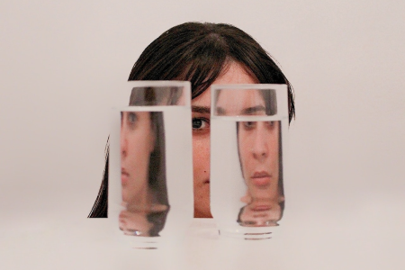 Image: a young woman's face is viewed through two glasses of water, so that the image is fractured into multiple angles.