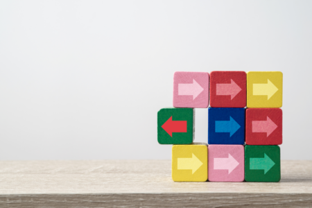Image: nine colorful wood blocks are stacked atop each other in a 3 by 3 square on a tabletop. Eight of the blocks are painted with a white arrow pointing to the right but one block, which is breaking away from the grid, is painted with a red arrow pointing to the left.