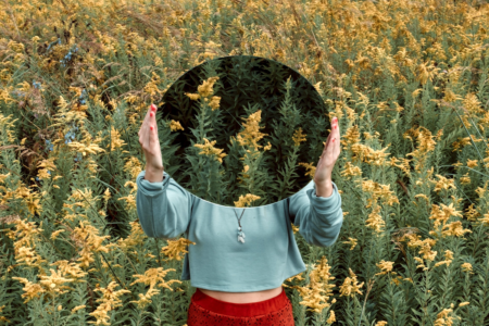 Image: a woman is standing in a field of wildflowers. She holds a round mirror in front of her so that, where we would otherwise see her head, we see only the reflection of wildflowers.