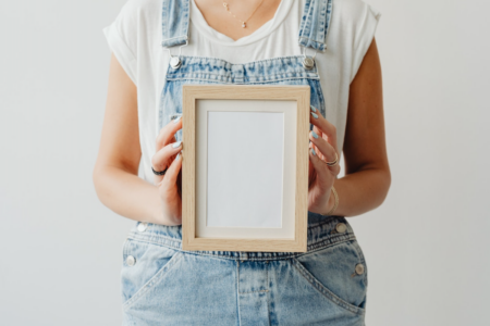 Image: a woman wearing blue overalls and a white t-shirt holds a small wooden picture frame to the viewer. The frame contains only empty white space where the picture should be.