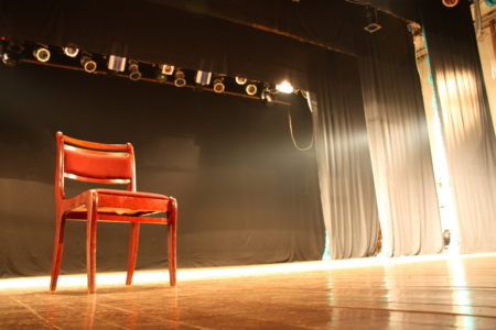 Image: a wooden chair sits alone on a brightly-lit theater stage.