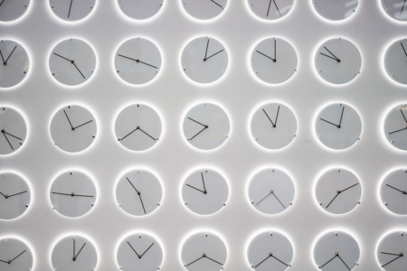 Image: on a white wall are mounted dozens of minimalist modern circular clocks which have minute and second hands all pointed in different directions, but no numbers.