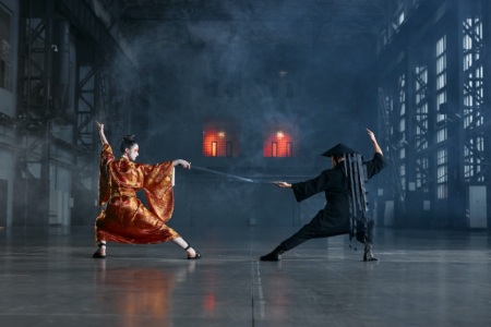 Image: in a large, smoky warehouse, a woman and man—she wearing an embroidered gold and red kimono, kabuki makeup and brandishing a sword, and he wearing a black samurai costume—face off for battle.