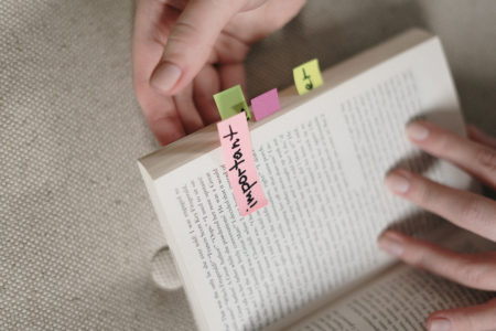 Image: a reader's hands hold a softcover book open. Some of the pages are flagged with sticky notes, including one on which the word "important" has been handwritten.