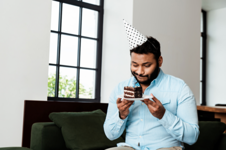 Image: a man wearing a conical paper party hat sits alone at home and considers eating a piece of cake, to illustrate the silver lining on a self-pity party.