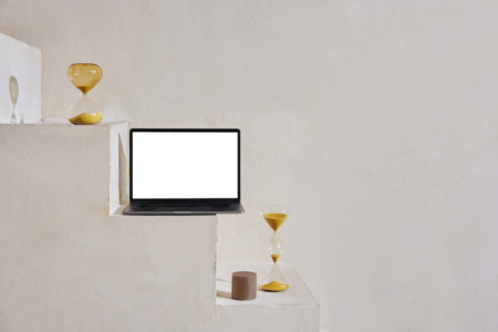 Image: an open laptop with a blank white screen sits on a step, flanked by decorative hourglasses.