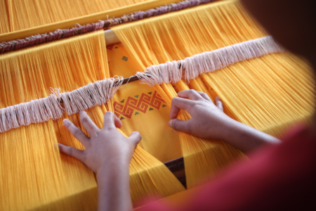 Image: a person at a weaving loom loaded with bright yellow thread pulls the threads apart to reveal that underneath is a finished version of the complex, multi-colored piece they're making, used for reference.