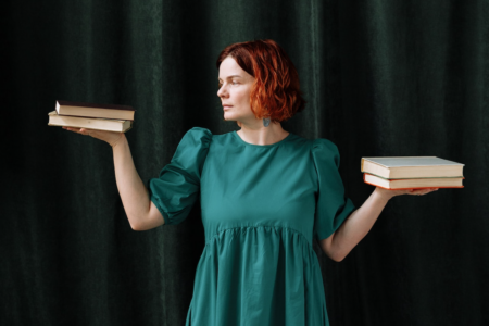 Image: a redheaded woman wearing a green dress stands in front of green curtains. In each upturned hand she holds two books, weighing them against one another in the manner of scales of justice.