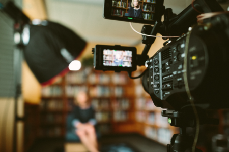 Image: in a room lined with bookshelves, a video camera and portable light are trained on a woman who's being interviewed.