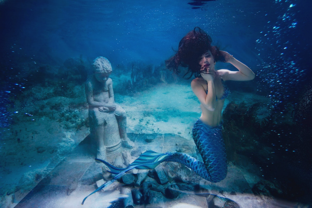 Image: a woman in a mermaid costume floats underwater next to sculptural ruins.