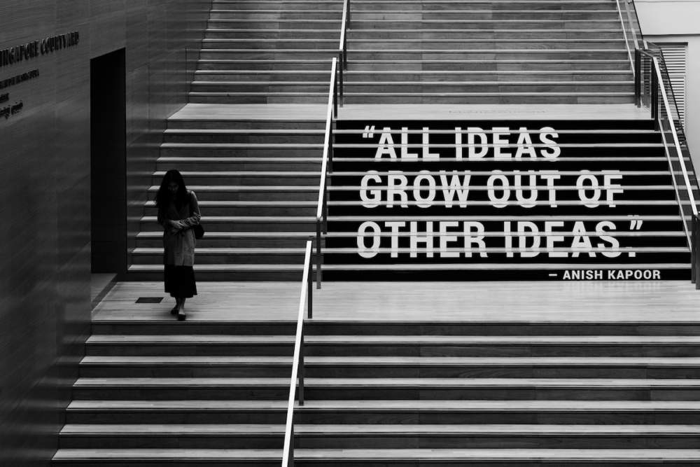 Image: a black and white photo of a woman walking down a massive indoor staircase on which a quote from Anish Kapoor is painted in large letters spanning many of the stair risers: "All ideas grow out of other ideas."