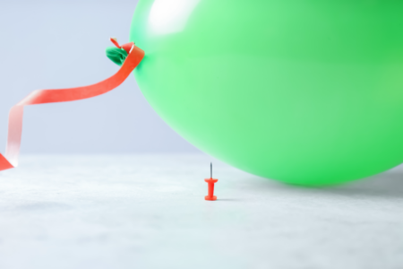 Image: a bright green balloon rests on a floor, hovering precariously over a red thumbtack with its pointed tip facing up.