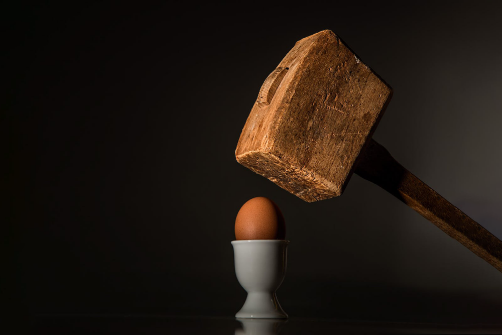 Image: a large, rough-hewn, wooden sledgehammer hovers above an egg resting in a ceramic egg cup.