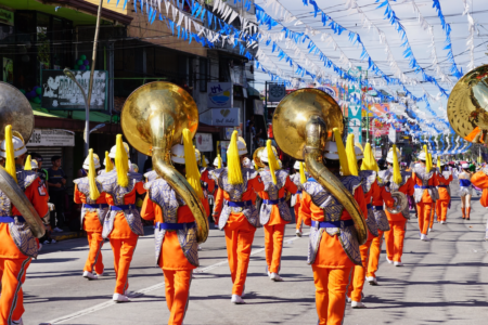 Image: a marching band in colorful costumes walks down an urban street, with blue and white bunting strung above.
