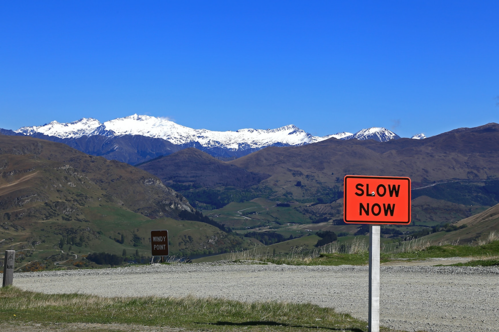 Image: against a dramatic backdrop of mountains, twisting roads, and a cloudless deep blue sky, a bright orange road sign reads "Slow Now".
