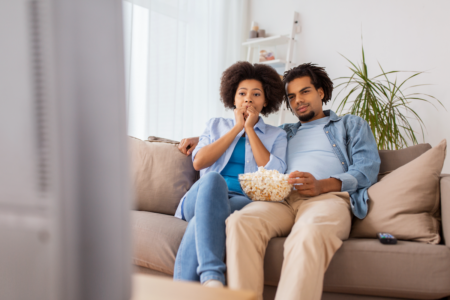 Image: a couple sits on the sofa, eating popcorn and watching television with rapt attention.