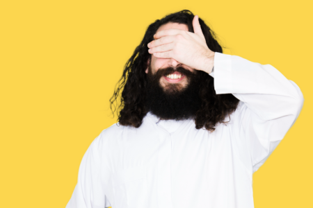 Image: a smiling man with long dark hair and a thick dark beard and wearing a white garment, as if meant to represent a Christ-like figure, covers his eyes with his hand.
