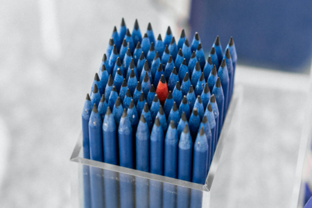 Image: a clear lucite box contains dozens of blue pencils standing vertically and tightly packed. At the center of them is a single red pencil.