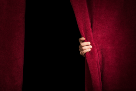 Image: the hand of an unseen person begins to pull back the heavy, red curtain of a theatrical stage as if they're about to emerge from the darkness.