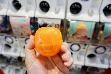 Image: against a backdrop of vending machines that dispense toys, an adult's hand holds an orange plastic bubble containing an unknown toy.