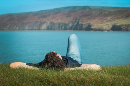 Image: a young man wearing jeans and a t-shirt reclines on the grass, facing the sky, near the water's edge a cloudless day.