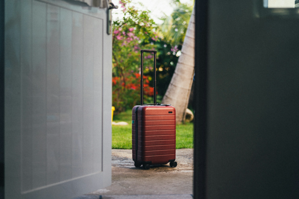 Image: Seen from inside the open doors of a room, a carry-on size suitcase with its handle up sits on an outdoor walkway against a backdrop of tropical foliage.