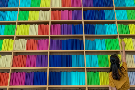 Image: a woman reaches up to grab a book from a set of shelves on which hundreds of books in solid-colored wrappers are arranged in rainbow stripes.