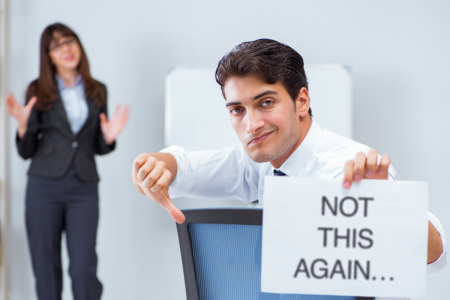 Image: in the background a woman in business attire is speaking. In the foreground a man who's turned to face the camera is giving a thumbs-down while his other hand holds a sign reading "Not this again…".