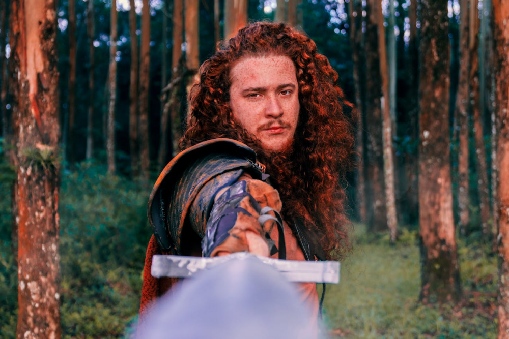Image: a man with long, curly, red hair and wearing colorful medieval garb stands in a forest and brandishes a sword at the viewer.
