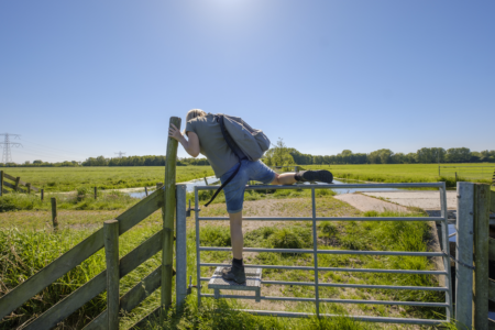 Image: a hiker wearing a backpack climbs over the locked gate of a fence in a rural area.