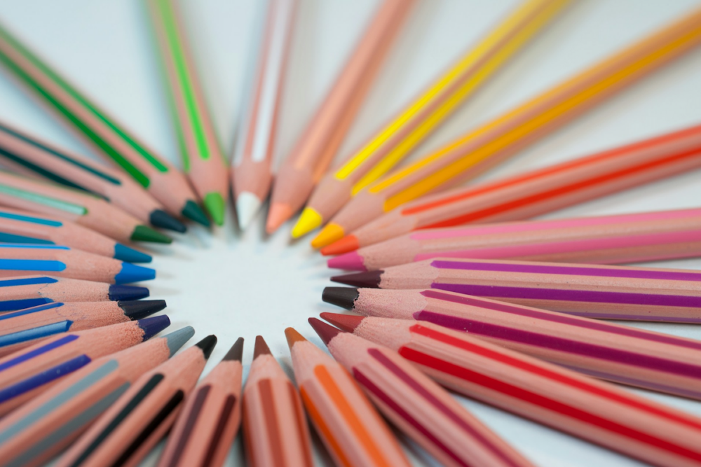 Image: like spokes of a wheel, colored pencils are arranged on a white table with their tips coming together in a multi-hued circle.