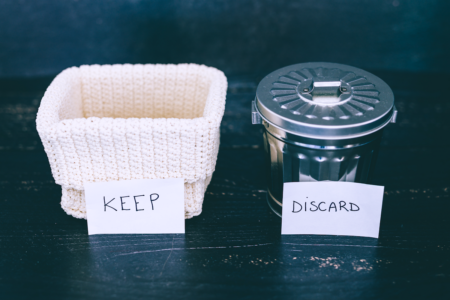 Image: on a black tabletop stand a small woven basket with a handmade sign reading "Keep" propped against it, next to a small metal garbage can with a sign reading "Discard" propped against it.