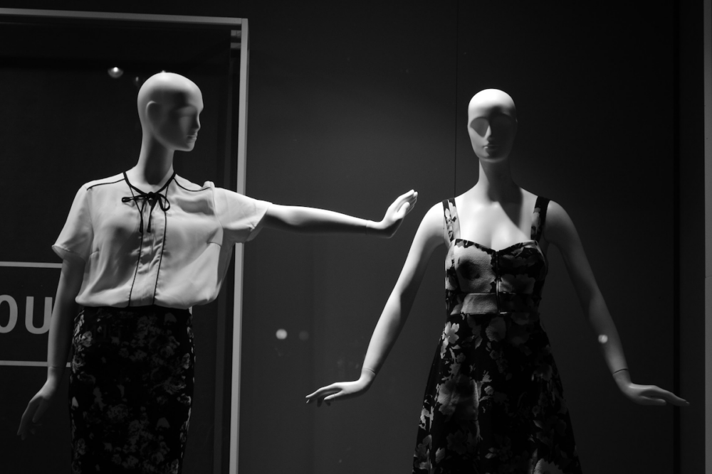 Image: black and white photo of two feminine mannequins in the window of a clothing store. One stares straight ahead with her arms at her sides, while the other's arm is outstretched and head is turned as if to make a connection with her companion.