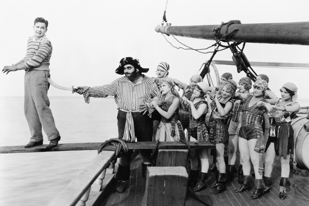 Image: in a black and white photo from what might be a an early motion picture, a young man in street clothes is being forced at sword point to walk the plank of a ship by a burly man in a pirate's costume and an all-woman crew.