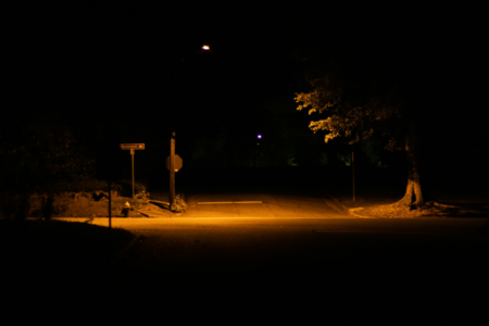 Image: a narrow swath of a suburban intersection is illuminated by a street lamp; otherwise everything is enveloped in darkness.