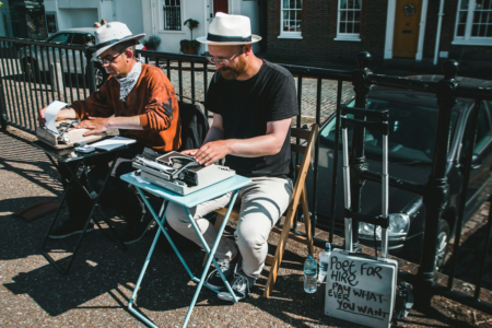Image: On an urban sidewalk, two men wearing casual clothing and hats sit side by side at folding tables on which typewriters are placed. Nearby they've placed a sign reading, "Poet for hire—pay whatever you want."