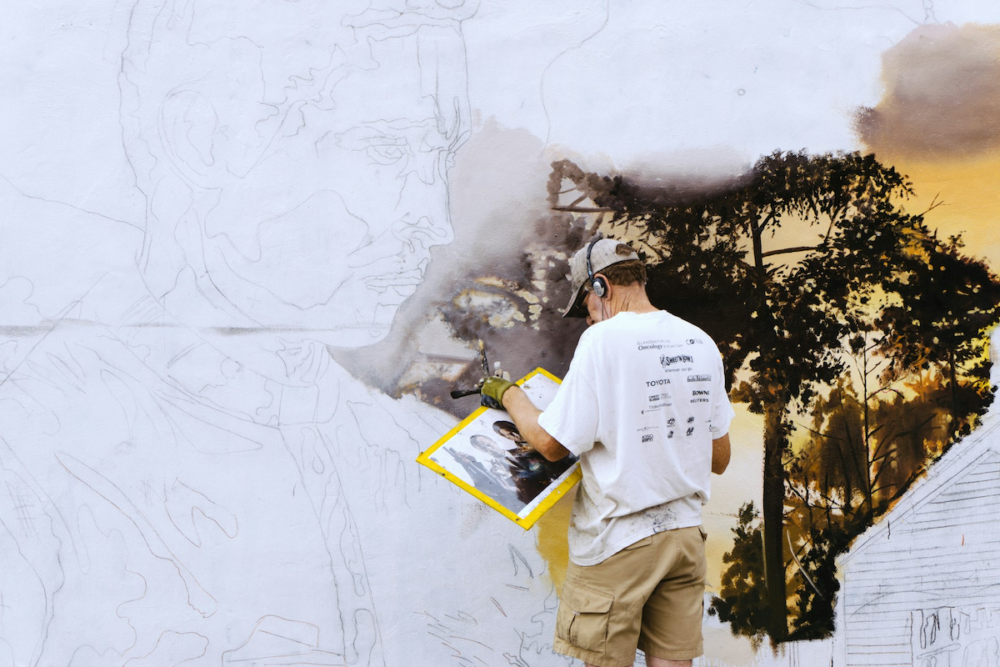 Image: a muralist has painted his image's background in great detail, while the main subject—a portrait which spans from top to bottom—remains only sketched in.