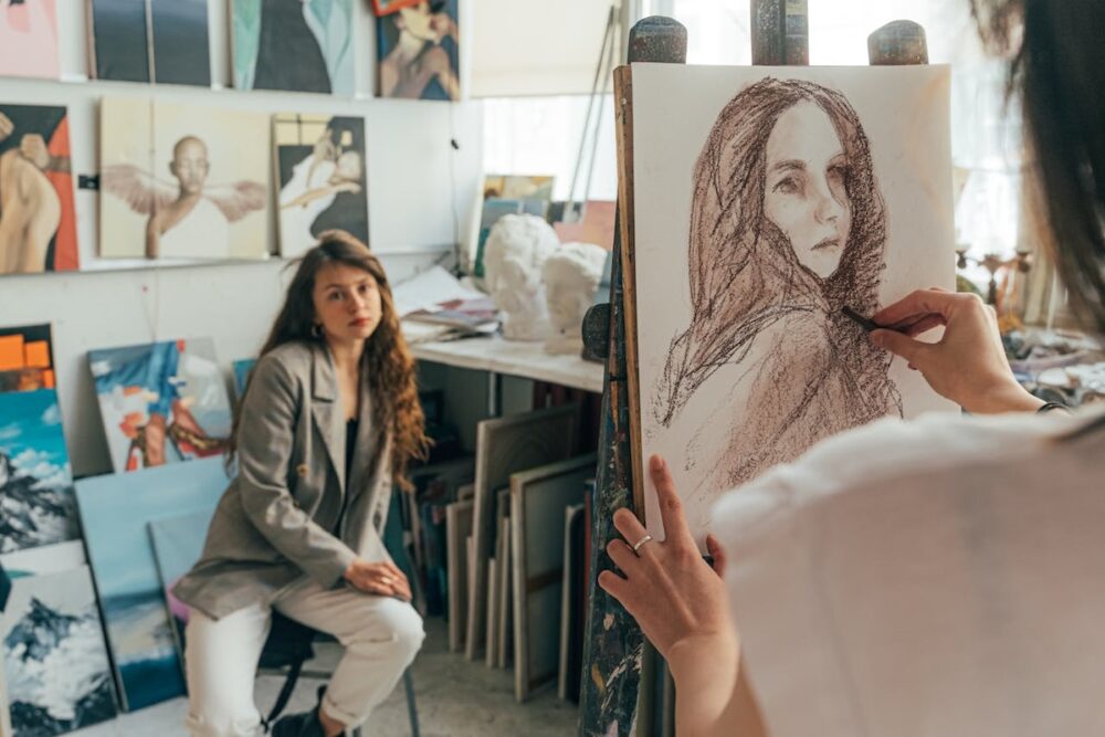 Image: In a light-filled studio an artist creates a charcoal portrait sketch of the model seated on a stool nearby.
